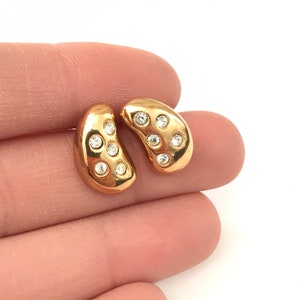 May include: A pair of gold-colored stud earrings. Each earring has a kidney bean shape and is adorned with several small, clear crystals. The earrings are displayed against a white background.
