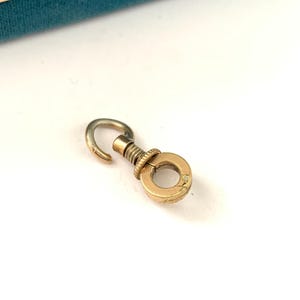 May include: A small, gold-toned metal clasp with a circular ring and a hook. The clasp has a screw-like design and is likely used for attaching items. The background is white.