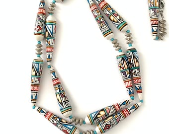 May include: A long necklace made of white ceramic beads with geometric patterns in shades of blue, red, yellow, and black. The beads are separated by silver spacers.