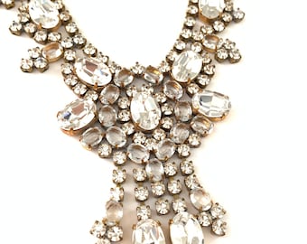 May include: A gold tone necklace with a large, teardrop-shaped crystal pendant. The pendant is surrounded by smaller crystals, creating a cascading effect. The necklace is made of metal and has a vintage look.