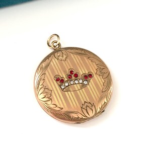 May include: A gold locket with a crown design featuring red and white stones. The locket has an engraved floral pattern and a striped background.