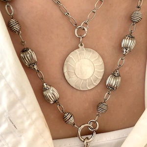 May include: A layered silver-toned necklace set featuring a shorter chain with a round, sunburst-patterned pendant and a longer chain adorned with alternating ribbed oval beads and textured spherical beads, finished with a toggle bar clasp.