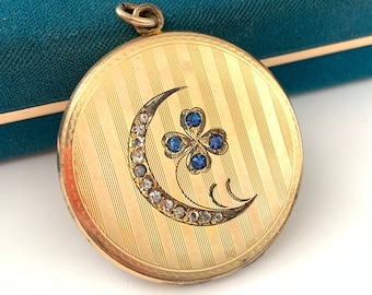 May include: A gold locket with a crescent moon design and a four-leaf clover with blue gemstones. The locket has a striped pattern.