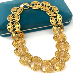 Op de afbeelding: Een gouden ketting met een geometrisch ontwerp. De ketting is gemaakt van in elkaar grijpende cirkels met een kleine cirkel in het midden van elke grotere cirkel.