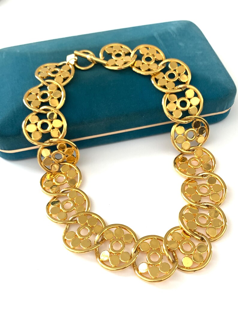 Op de afbeelding: Een gouden ketting met een geometrisch ontwerp. De ketting is gemaakt van in elkaar grijpende cirkels met een kleine cirkel in het midden van elke grotere cirkel.