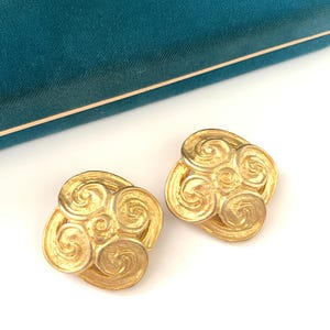 May include: A pair of gold-toned, square-shaped clip-on earrings. Each earring features a swirling, scroll-like design. The earrings are set against a teal velvet jewelry box.