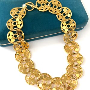 Op de afbeelding: Een gouden ketting met een ketting van in elkaar grijpende cirkels. Elke cirkel heeft een kleinere cirkel die uit het midden is geknipt, waardoor een delicaat en ingewikkeld ontwerp ontstaat.