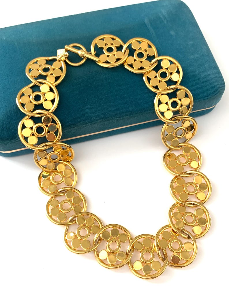 Op de afbeelding: Een gouden ketting met een ketting van in elkaar grijpende cirkels. Elke cirkel heeft een kleinere cirkel die uit het midden is geknipt, waardoor een delicaat en ingewikkeld ontwerp ontstaat.