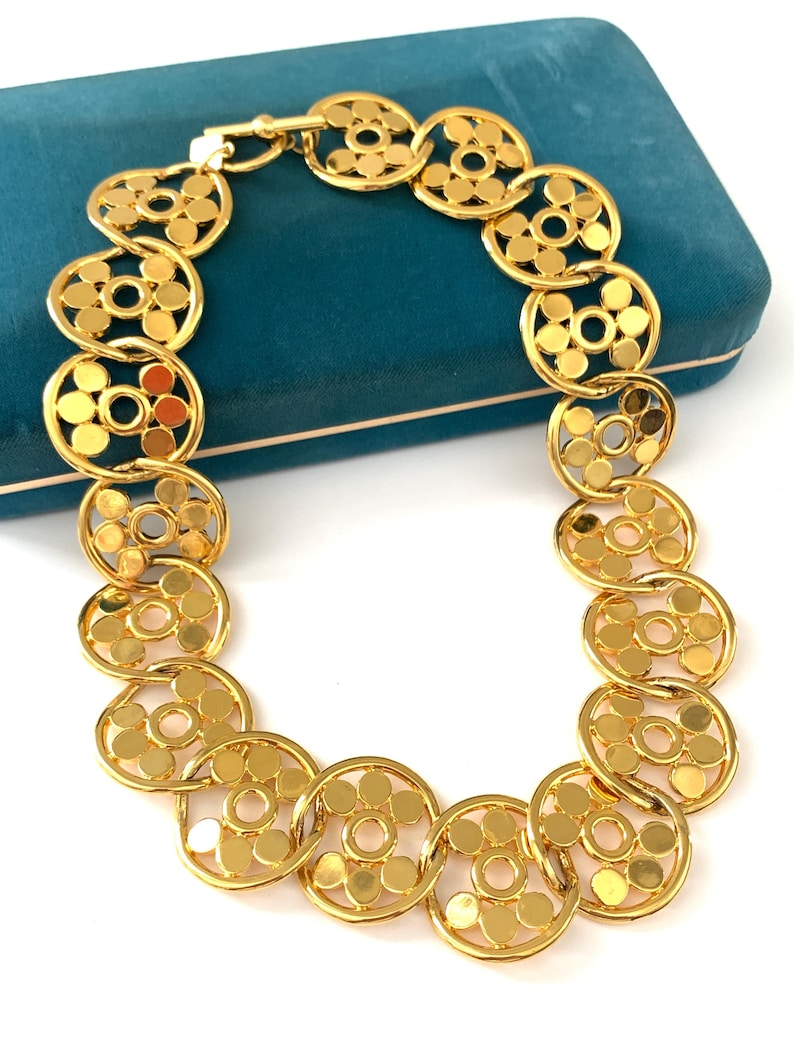 Op de afbeelding: Een gouden ketting met een geometrisch ontwerp. De ketting is gemaakt van in elkaar grijpende cirkels met een bloemmotief.
