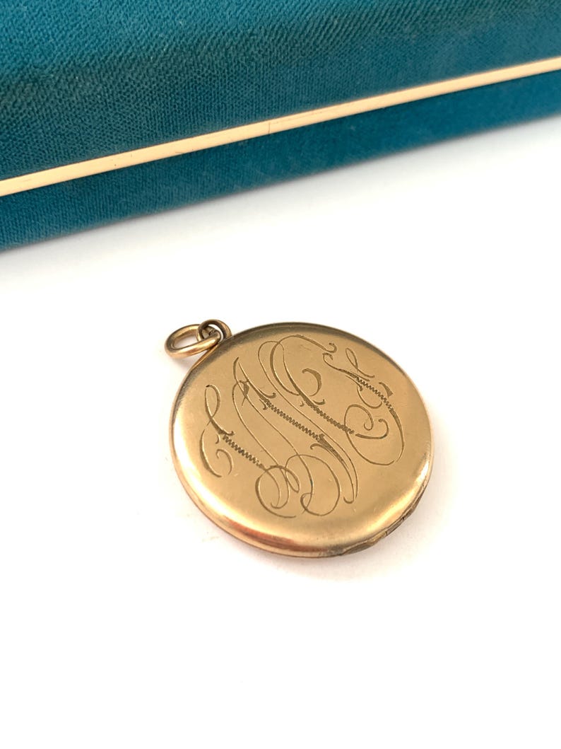 Puede incluir: Medall&oacute;n de oro con un monograma grabado. El medall&oacute;n es redondo y tiene una tapa con bisagras. El monograma est&aacute; en una escritura cursiva.