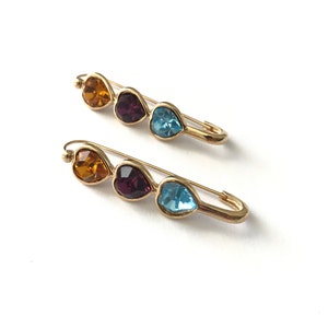 Pu&ograve; includere: Due spille di sicurezza dorate con tre strass a forma di cuore. Gli strass sono arancioni, viola e blu.