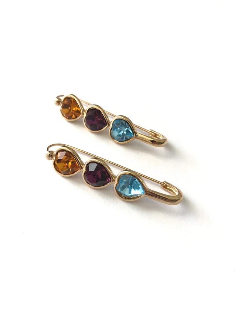 Pu&ograve; includere: Due spille di sicurezza dorate con tre strass a forma di cuore. Gli strass sono arancioni, viola e blu.