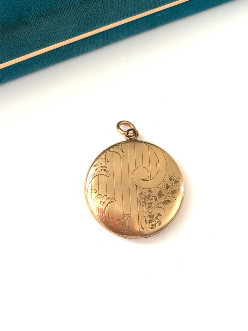 Puede incluir: Un medall&oacute;n ovalado dorado con un dise&ntilde;o floral grabado. El medall&oacute;n tiene una cadena delicada unida a la parte superior.