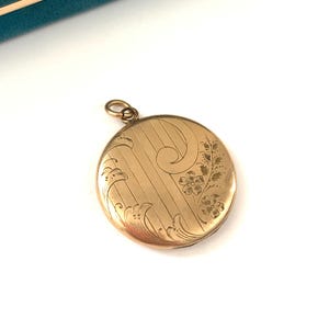 Puede incluir: Un medall&oacute;n redondo dorado con un dise&ntilde;o floral grabado. El medall&oacute;n tiene una cadena delicada unida a la parte superior.