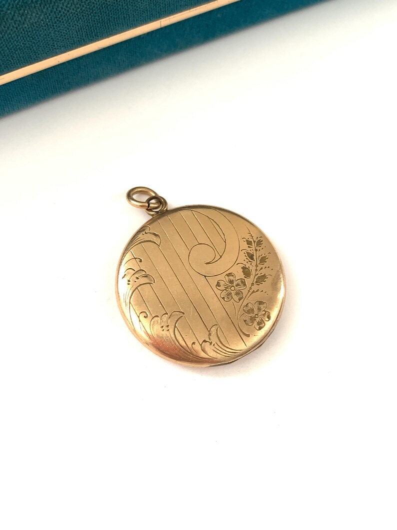 Puede incluir: Un medall&oacute;n redondo dorado con un dise&ntilde;o floral grabado. El medall&oacute;n tiene una cadena delicada unida a la parte superior.