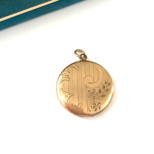 Puede incluir: Un medall&oacute;n redondo de color dorado con un dise&ntilde;o floral grabado. El medall&oacute;n es redondo y tiene una cadena delicada unida a la parte superior.