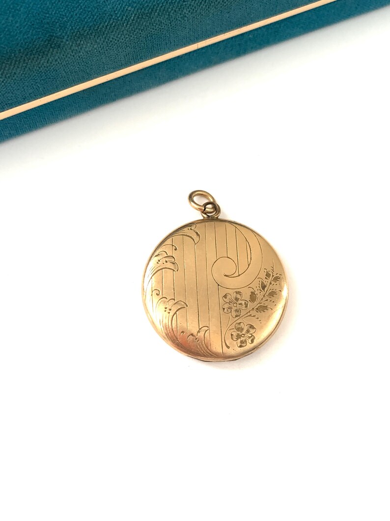 Puede incluir: Un medall&oacute;n redondo de color dorado con un dise&ntilde;o floral grabado. El medall&oacute;n es redondo y tiene una cadena delicada unida a la parte superior.