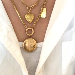Puede incluir: Tres collares de oro con diferentes colgantes. El collar superior tiene un peque&ntilde;o colgante rectangular en forma de placa. El collar del medio tiene un colgante en forma de coraz&oacute;n. El collar inferior tiene un colgante redondo con dise&ntilde;os florales.
