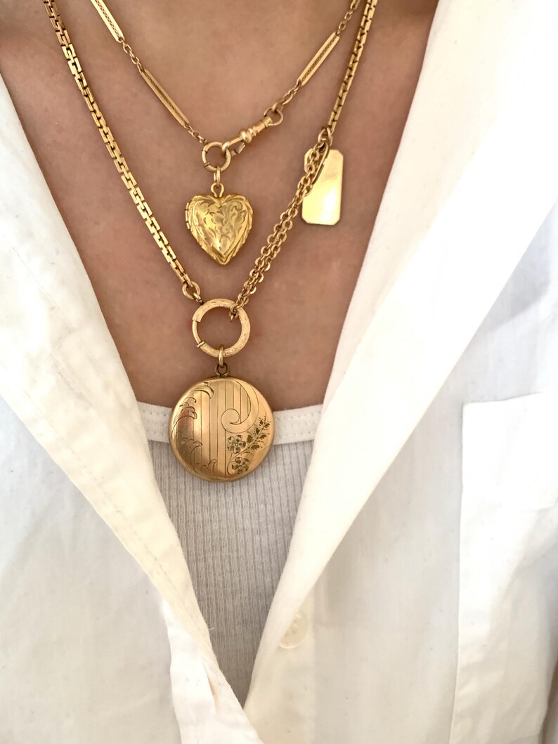 Puede incluir: Tres collares de oro con diferentes colgantes. El collar superior tiene un peque&ntilde;o colgante rectangular en forma de placa. El collar del medio tiene un colgante en forma de coraz&oacute;n. El collar inferior tiene un colgante redondo con dise&ntilde;os florales.