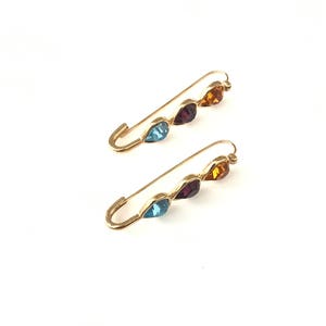 Pu&ograve; includere: Due spille di sicurezza dorate con tre strass a forma di goccia ciascuna. Gli strass sono blu, viola e arancioni.