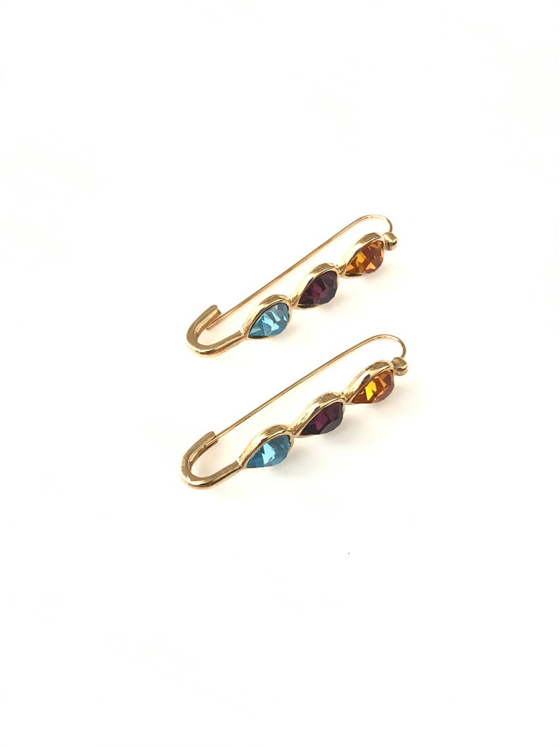 Pu&ograve; includere: Due spille di sicurezza dorate con tre strass a forma di goccia ciascuna. Gli strass sono blu, viola e arancioni.