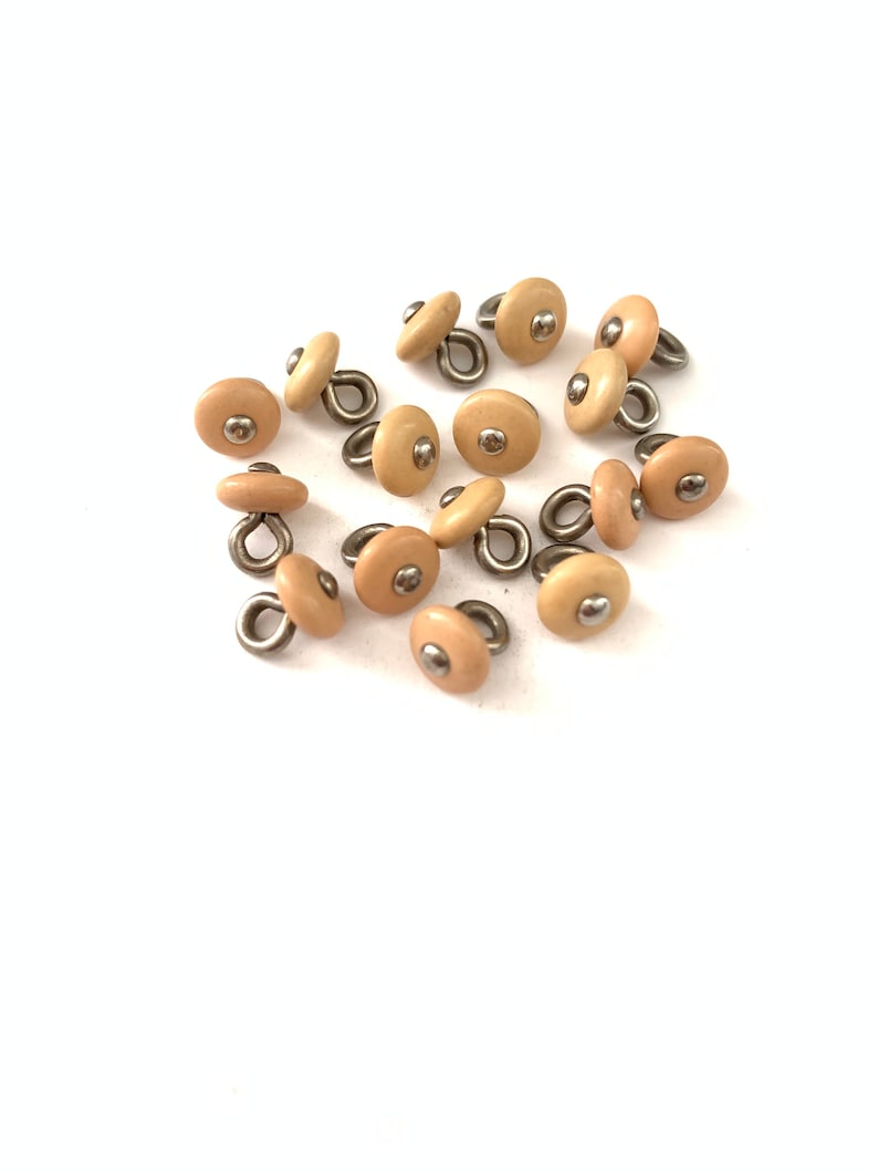 May include: A group of 14 beige plastic buttons with a metal loop on the back. The buttons have a small silver dot in the center.