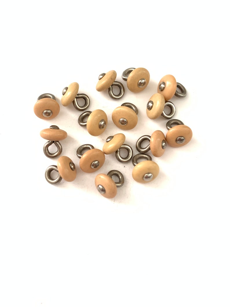 May include: A group of 15 beige plastic buttons with a silver loop and a silver dot in the center.