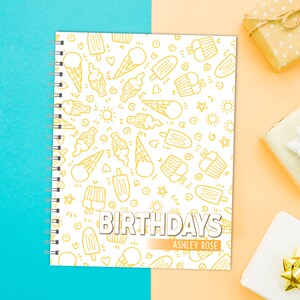 May include: A white spiral bound notebook with a yellow ice cream cone and popsicle pattern. The text "BIRTHDAYS" is printed in yellow on the cover with the name "ASHLEY ROSE" printed below.