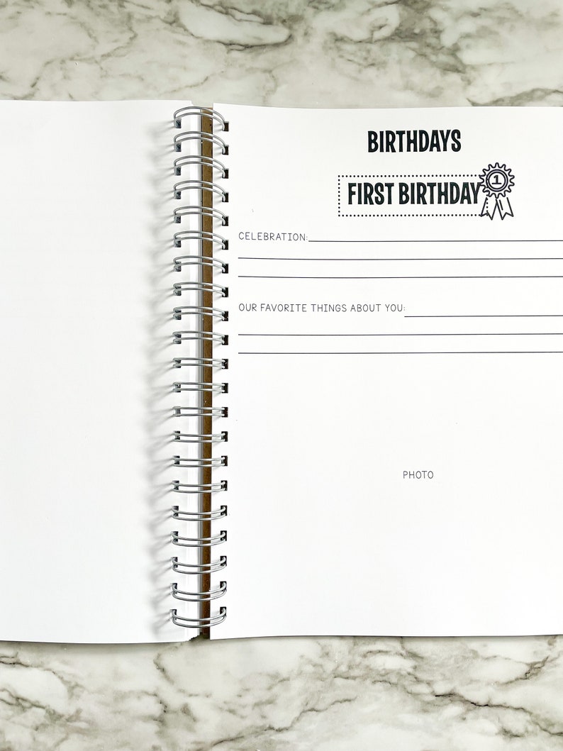 K&ouml;nnte beinhalten: Eine wei&szlig;e Seite aus einem Erinnerungsbuch zum ersten Geburtstag eines Babys mit dem Text "BIRTHDAYS" und "FIRST BIRTHDAY" in Schwarz. Die Seite hat Zeilen zum Schreiben &uuml;ber die Feier und die Lieblingsdinge des Babys. Es gibt Platz f&uuml;r ein Foto.