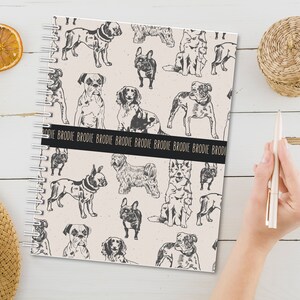 May include: A cream-colored spiral bound notebook with a black and white illustration of various dog breeds. The notebook has a black band with the repeated text "Brodie" in gold lettering.