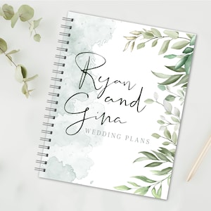 May include: A white wedding planner with a watercolor background and green foliage. The text "Ryan and Gina Wedding Plans" is printed on the front.