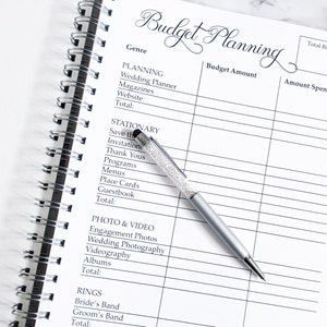 May include: A white budget planner with a black spiral binding. The planner has columns for genre, budget amount, and amount spent. The planner is open to a page with categories for planning, stationary, photo and video, and rings.
