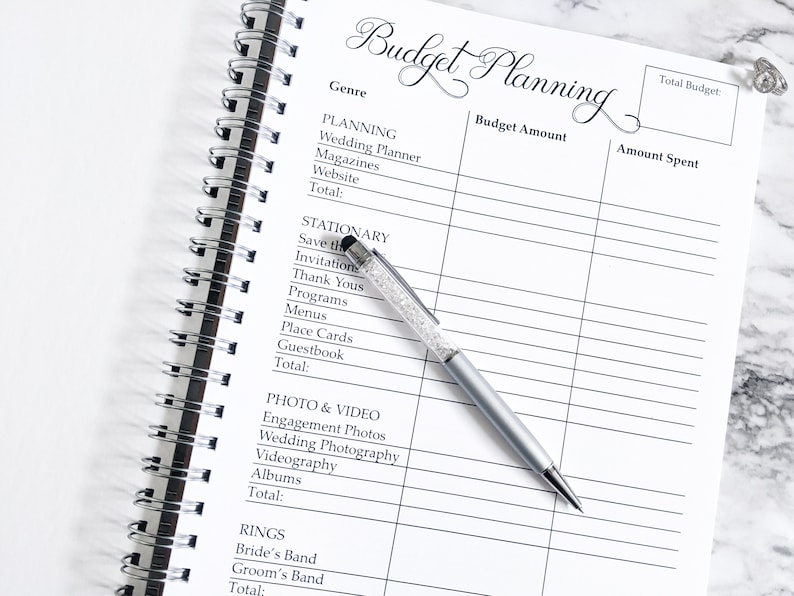 May include: A white budget planner with a black spiral binding. The planner has columns for genre, budget amount, and amount spent. The planner is open to a page with categories for planning, stationary, photo and video, and rings.