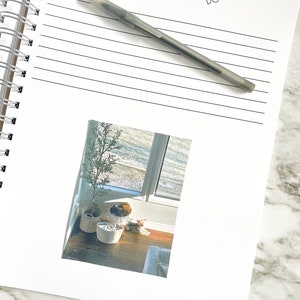 K&ouml;nnte beinhalten: Ein wei&szlig;es Notizbuch mit dem Wort "MEMORIES" und einem Pfotenabdruck in schwarzer Tinte. Ein Stift liegt auf dem ge&ouml;ffneten Notizbuch. Ein Foto eines Hundes und einer Katze, die vor einem Fenster sitzen, ist an das Notizbuch befestigt.