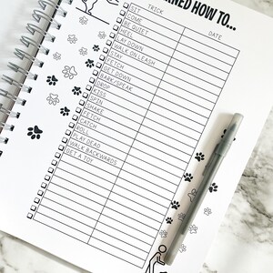 K&ouml;nnte beinhalten: Eine schwarz-wei&szlig;e Checkliste mit Pfotenabdr&uuml;cken an der Seite. Die Checkliste tr&auml;gt den Titel "Ich habe gelernt, wie man..." und enth&auml;lt eine Liste von Hundetricks mit Kontrollk&auml;stchen neben jedem Trick. Die Tricks umfassen Sitz, Komm, Sei ruhig, Fu&szlig;, Leg dich hin, Geh an der Leine, Bleib, Hol, Geh runter, Bell/Sprich, Lass fallen, K&uuml;ss, Dreh dich, Sch&uuml;ttel, Hol, Fang, Roll dich, Spiel tot, Geh r&uuml;ckw&auml;rts und Hol dir ein Spielzeug.