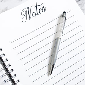 May include: A white spiral bound notebook with the word "Notes" written in black cursive at the top. A silver pen with a clear crystal design and a black stylus tip is lying on the notebook.