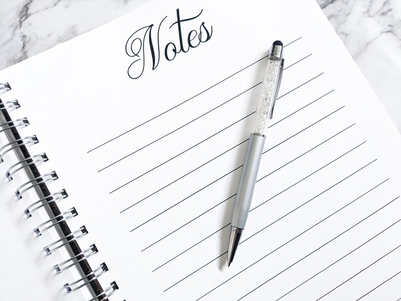 May include: A white spiral bound notebook with the word "Notes" written in black cursive at the top. A silver pen with a clear crystal design and a black stylus tip is lying on the notebook.