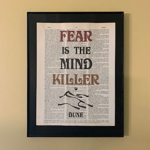 May include: A framed print of a dictionary page with the words "Fear is the mind killer" in large, bold letters. The words are printed in brown and black on a white background. The print is framed in black.