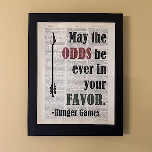 May include: A black and white print of a dictionary page with the quote "May the odds be ever in your favor - Hunger Games" in red, black, and green text. An arrow is printed in black on the left side of the page.