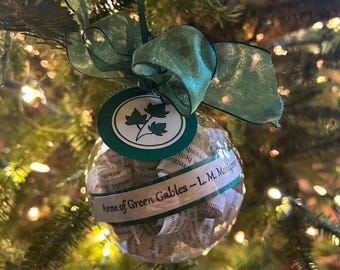 May include: A clear glass Christmas ornament filled with rolled up pages from the book "Anne of Green Gables" by L.M. Montgomery. The ornament is decorated with a green ribbon and a green and white tag with a leaf design.