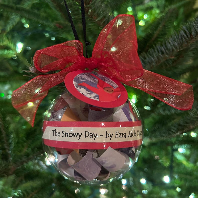 May include: A clear glass ornament with a red ribbon and a tag that says "The Snowy Day - by Ezra Jack Keats". The ornament is filled with small pieces of paper.