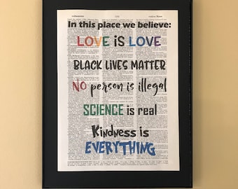May include: A framed print with a dictionary page background and the text "In this place we believe: LOVE IS LOVE, BLACK LIVES MATTER, No person is illegal, SCIENCE is real, Kindness is EVERYTHING" in colorful lettering.