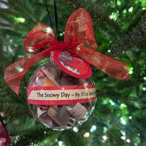 May include: A clear glass Christmas ornament with a red ribbon and a red and white paper circle with the text "The Snowy Day - by Ezra Jack Keats". The ornament is filled with small pieces of paper.