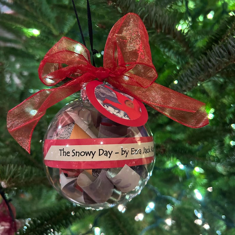 May include: A clear glass Christmas ornament with a red ribbon and a red and white paper circle with the text "The Snowy Day - by Ezra Jack Keats". The ornament is filled with small pieces of paper.