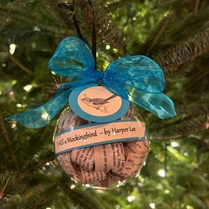 May include: A clear glass Christmas ornament filled with rolled up pages from the book "To Kill a Mockingbird" by Harper Lee. The ornament is decorated with a blue ribbon and a tag with a small bird illustration.