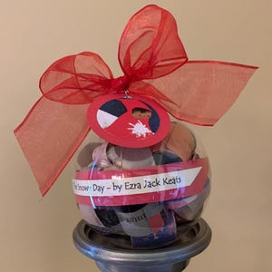 May include: A clear glass ornament filled with colorful paper scraps and a red ribbon. The ornament has a red tag with a black and white illustration of a child in a red coat. The tag also has the text "The Snow Day - by Ezra Jack Keats".