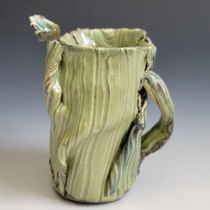 May include: A ceramic pitcher with a light green glaze and a textured, vertical ribbed surface. The vessel features an irregular, organic rim, a prominent spout on the left side, and a curved handle on the right, all with dark brown accents.