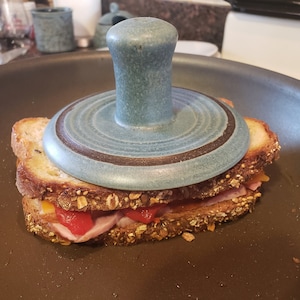 May include: A grilled sandwich being pressed with a blue ceramic sandwich press. The sandwich is made with toasted bread, ham, cheese, and red peppers. The sandwich press is round with a handle.