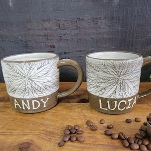 May include: Two ceramic mugs with brown and white leafy designs. The mugs have the names "Andy" and "Lucia" written on the bottom in white lettering.