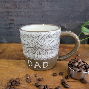 May include: A white ceramic mug with a brown handle and a leafy design. The bottom of the mug is brown and has the word "DAD" written on it. The mug is sitting on a wooden surface with coffee beans scattered around it.